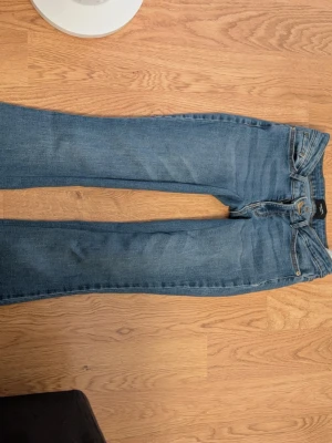 Blå lätt utsvängda jeans med klassisk passform - Säljer ett par blå jeans med rak passform och låg midja. Jeansen har fem fickor, bälteshällor och stängs med dragkedja och knapp. Materialet är klassisk denim i bomull. Perfekta till en avslappnad stil.