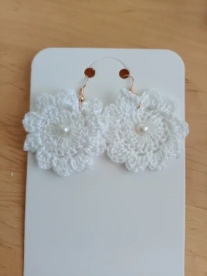 Vita virkade blomörhängen med pärla - Unika örhängen i form av vita virkade blommor med en liten pärla i mitten. Hängena har guldfärgade krokar och ger en söt och bohemisk känsla. Perfekta för dig som vill ha något handgjort och personligt.