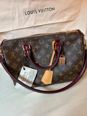 Speedy 30 Soft Celebration  - Säljer denna superfina och unika speedy 30 i den otroligt eftertraktade ”Soft Celebration”-modellen med vinröda/bordeaux läderdetaljer. Den perfekta vardagsväskan som rymmer allt du behöver och kan bäras både i handtagen eller som crossbody/axelväska tack vare den medföljande remmen.Väskan är köpt "preloved" via lyxplattformen Vestiaire Collective. Jag fick ej butiks kvittot eller något separat äkthetsintyg, och jag kan därför inte garantera äktheten personligen