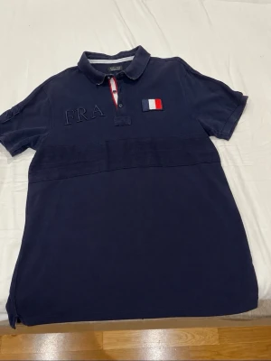 Marinblå pikétröja från Zara Man - Snygg marinblå pikétröja från Zara Man med broderad FRA-text och fransk flagga på bröstet. Tröjan har klassisk krage, korta ärmar och knappar framtill med rödvit detalj. Perfekt för en sportig och stilren look.