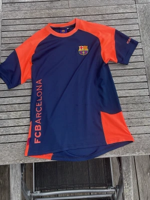FC Barcelona träningströja - Snygg FC Barcelona träningströja i marinblått med rosa/orange detaljer och klubbmärke på bröstet. Tröjan har korta ärmar, rund hals och är tillverkad i ett lätt, ventilerande material som passar perfekt till träning eller sport.