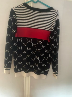 Svart stickad tröja från Gucci - Snygg svart stickad tröja från Gucci med vita GG-loggor över hela plagget. Överdelen har vita och svarta ränder samt en bred röd panel över bröstet. Tröjan har långa ärmar och vita ribbade muddar vid ärmslut och nederkant. Perfekt för dig som gillar statement-plagg.