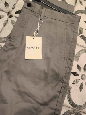 Grå chinos från GANT - Snygga grå chinos från GANT med klassisk passform och raka ben. Byxorna har sidofickor och bälteshällor, perfekta för en clean och stilren look. Tillverkade i bomull.