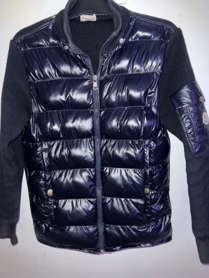 Moncler cardigan  - Snygg mörkblå cardigan med glansig framsida och stickade ärmar samt rygg. Jackan har dragkedja framtill, två fickor med tryckknappar och en patch på ena ärmen. Perfekt för lager-på-lager och streetstyle.