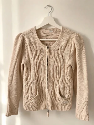Beige stickad kofta från Odd Molly - Säljer en beige stickad kofta från Odd Molly med dragkedja framtill och snygga flätstickade detaljer. Koftan har rund halsringning, långa ärmar och ribbade muddar. Perfekt för lager på lager och ger en mysig känsla till din outfit.