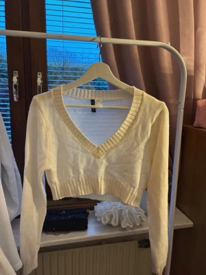 Vit croppad stickad tröja V-ringad - Säljer en vit croppad stickad tröja med djup V-ringning och långa ärmar. Tröjan har ribbade muddar vid ärmslut och nederkant samt en bred ribbad kant runt halsen. Perfekt att styla med höga jeans eller kjol.🌸