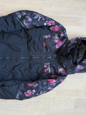 Svart vindjacka med blommigt mönster. Stl. 134  - Snygg svart vinter-/skidjacka med blommigt mönster i rosa och lila på ärmar och huva. Jackan har dragkedja framtill, fickor och en justerbar huva. .