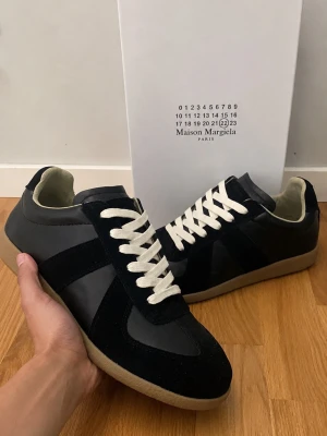 Maison Margiela svarta sneakers - Maison Margiela sneakers i svart läder och mocka med vita skosnören och beige gummisula. Klassisk låg modell med stilrena detaljer och diskret logotyp på baksidan. Perfekta för dig som gillar minimalistisk och tidlös design.