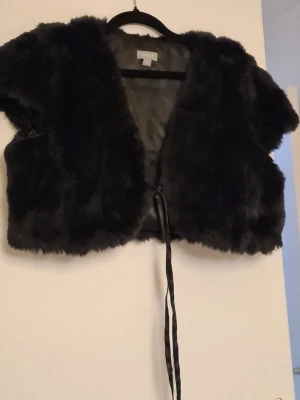 Kort svart fuskpälsväst med knytband - Snygg kort svart fuskpälsväst med mjukt foder och knytband framtill. Västen har en öppen design utan krage och är perfekt för att lyfta en outfit. Passar dig som gillar trendiga och mysiga plagg med lite extra attityd.