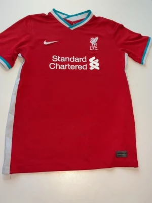 Liverpool Nike matchtröja röd - Säljer en röd Liverpool matchtröja från Nike med vit och turkos detalj vid krage och ärmslut. Tröjan har klubbmärke och sponsortryck framtill samt Dri-FIT-material som andas. Perfekt för dig som älskar fotboll och vill bära ditt lag med stil. 