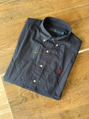 Mörkblå/Svart skjorta från Ralph Lauren - Klassisk mörkblå skjorta från Ralph Lauren med knappslå och vit knappstängning. Skjortan har lång ärm och ikonisk röd logga broderad på bröstet. Tillverkad i mjuk bomull och har en tidlös, normal passform.
