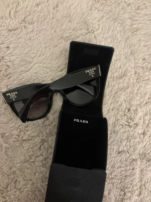Svarta solglasögon från Prada - Snygga svarta solglasögon från Prada med breda skalmar och klassisk design. Glasen är mörka och bågarna har tydlig Prada-logga på sidan. Kommer med originalfodral. Perfekta för att lyfta din stil till nästa nivå.