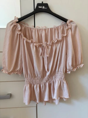 Ljusrosa offshoulder blus med volang - Supersöt ljusrosa blus med offshoulder-design, puffiga korta ärmar och volangkant. Smockad midja ger en snygg passform och knytband framtill. Perfekt för dig som vill ha en romantisk och trendig topp till sommaren.