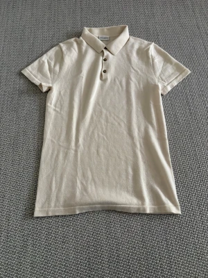 Beige pikétröja från John Henric - Säljer en stilren beige pikétröja från John Henric med klassisk krage och tre knappar framtill. Tröjan är i mjukt material och har korta ärmar, perfekt för en clean och enkel look.