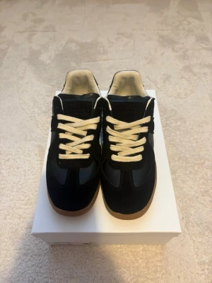 Maison Margiela GAT - strl 43  - Stilrena svarta sneakers med kontrasterande vita skosnören och ljusbrun gummisula. Ovandelen är i en mix av mocka och slätt läder, vilket ger en snygg och modern look. Perfekta för dig som gillar klassisk men trendig stil.