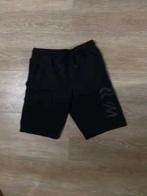 Svarta shorts med snörning - Svarta shorts med snörning i midjan och tryckt text på ena benet. Modellen har en avslappnad passform och är perfekta för en chill stil. Shortsen har fickor på sidorna och är tillverkade i ett mjukt material.