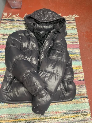 Svart pufferjacka från Brave Soul - Snygg svart pufferjacka från Brave Soul med glansig finish och huva. Jackan har dragkedja framtill, två sidofickor med dragkedja och en extra ficka på bröstet. Perfekt för kalla vinterdagar och riktigt skön att bära.