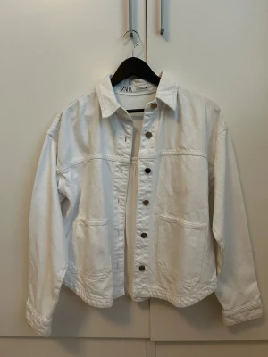 Vit jeansjacka från Zara - Snygg vit jeansjacka från Zara med raka linjer och avslappnad passform. Jackan har stora framfickor, klassisk krage och metallknappar framtill. Perfekt att slänga över en hoodie eller t-shirt för en clean och fräsch look.