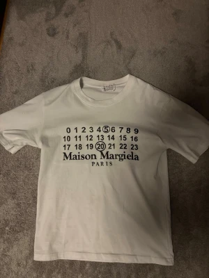 Maison Margiela vit t-shirt - Vit t-shirt från Maison Margiela med klassiskt tryck framtill med siffror och logga. Rund halsringning och korta ärmar. Perfekt för dig som gillar minimalistisk och stilren design.