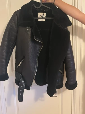 Svart bikerinspirerad kappa från ZARA - Snygg svart kappa från ZARA i bikerstil med fuskpälsfoder och bred krage. Jackan har dragkedjor, bälte i midjan och detaljerade sömmar. Perfekt för kalla dagar och ger en cool look till vilken outfit som helst.