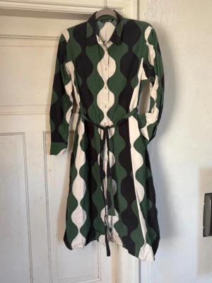 Grön och svart Marimekko klänning - Säljer en unik Marimekko x Uniqlo klänning i grönt, svart och vitt med vågigt mönster. Klänningen har lång ärm, knappar framtill och ett matchande bälte i midjan. Perfekt för dig som gillar statement-plagg och grafiska prints. Fickor i sidorna 