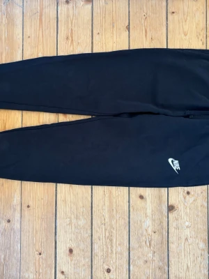 Svarta mjukisbyxor från Nike i bomull storlek XS - Säljer ett par svarta mjukisbyxor från Nike med klassisk logga på benet. Byxorna har avsmalnande ben och resår i midjan för skön passform. Materialet är bomull. Storlek XS