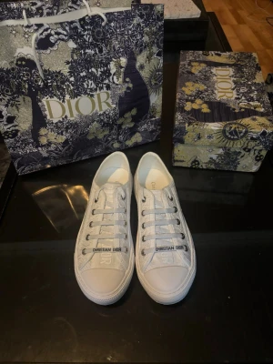 Vita Dior sneakers med logga - Säljer ett par vita Dior sneakers med tydlig Christian Dior-logga på snörningen. Skorna har lågt skaft och är tillverkade i canvas med subtilt mönster i vitt och grått. Klassisk siluett med vita snören och gummisula. Perfekta för dig som gillar exklusiv streetstyle.
