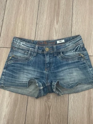 Blå jeansshorts med slitningar (pris kan diskuteras!) - Säljer ett par blå jeansshorts med slitna detaljer och råa kanter. Shortsen har fem fickor, nitar och en knappstängning framtill. Perfekta för varma dagar och ger en avslappnad look.