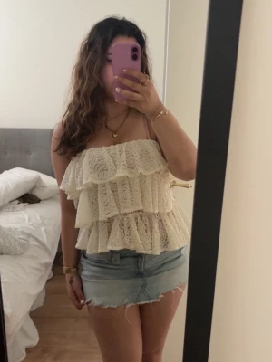 Vit spetsig volanglinnetopp - Supersöt vit linnetopp med flera lager volanger och spetsdetaljer. Axelbandslös design med romantisk känsla, perfekt för varma dagar. Toppen har en luftig och feminin look som passar till jeansshorts eller kjol.