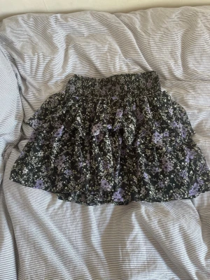 Svart blommig volangkjol - Kort svart kjol med lila och vita blommor och volangdetaljer. Kjolen har resår i midjan och ett luftigt, flowy tyg som passar perfekt till varmare dagar. 