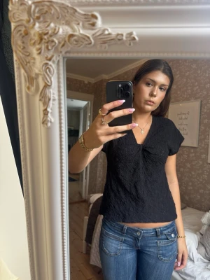 Svart v-ringad topp med struktur - Säljer en svart topp med v-ringning och korta ärmar. Tyget har en snygg strukturerad yta som ger en unik look. Passformen är normal och toppen är lätt croppad, perfekt till jeans eller kjol.