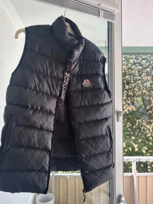 Svart dunväst från Moncler - Snygg svart dunväst från Moncler med hög krage och dragkedja framtill. Västen har Moncler-logga på bröstet och bred dragkedja med broderad text. Perfekt för lager-på-lager och håller dig varm under kyliga dagar.
