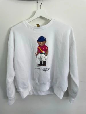 Vit sweatshirt Polo Bear Ralph Lauren - Aldrig använd! (Julklapp) Vit sweatshirt med Polo Bear-tryck från Ralph Lauren. Tröjan har rund halsringning, långa ärmar och ribbade muddar. Trycket visar en björn i röd pikétröja och blå hjälm. Perfekt för en avslappnad och trendig stil.