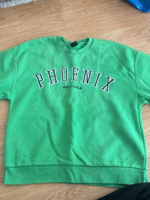 Grön sweatshirt med PHOENIX-tryck - Snygg grön sweatshirt med texten PHOENIX ARIZONA tryckt på bröstet. Tröjan har rund halsringning, ribbade muddar vid ärmslut och nederkant samt en avslappnad passform. Perfekt för en chill och sportig look.