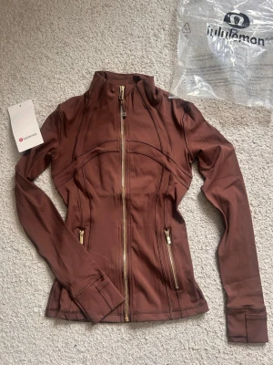  Lululemon size 2 - Slimmad brun träningsjacka från Lululemon med guldfärgade dragkedjor och två fickor framtill. Tillverkad i ett stretchigt funktionsmaterial som passar perfekt till träning eller som ett extra lager. Snygga sömdetaljer och hög krage för sportig look.