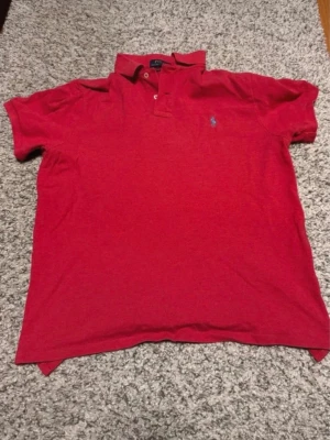 Röd pikétröja från Ralph Lauren - Klassisk röd pikétröja från Ralph Lauren med blå broderad logga på bröstet. Tröjan har korta ärmar, krage och knappar vid halsen. Tillverkad i mjuk bomull som känns skön mot huden. Perfekt för en avslappnad men ändå stilren look. L i barnstorlek