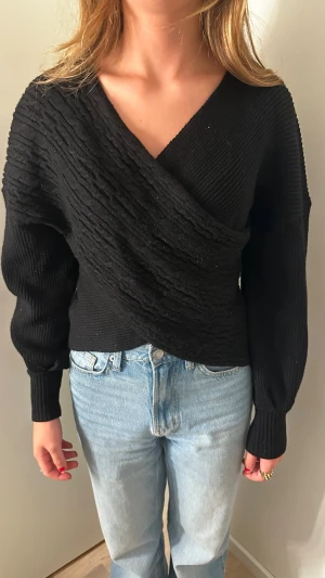 Svart omlottstickad tröja - Snygg svart stickad tröja med omlott-design och strukturstickade detaljer framtill. Tröjan har långa ärmar med ribbade muddar och en djup v-ringning. Perfekt till jeans för en avslappnad men trendig look.