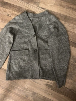Grå stickad kofta med fickor - Mysig grå stickad kofta med två stora ribbade fickor framtill och knäppning med knappar. Koftan har bred ribbad mudd vid ärmslut och nederkant samt en djup V-ringning. Perfekt att slänga på sig när det är kyligt ute.