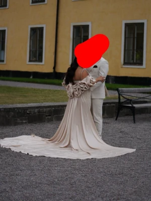 Champagnefärgad klänning med pärlor - Elegant champagnefärgad klänning med smala axelband och lång släp. Klänningen har ett lager av pärlor framtill och en avtagbar släp med stora rosetter. Köptes för 8000 säljes för 5000. Storlek M. Den kommer behövas att kemtävttas.