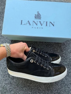 Svarta Lanvin sneakers med lackdetaljer - Snygga svarta sneakers från Lanvin med ovandel i mocka och lackad tå. Skorna har vita sulor, svarta skosnören och metallfärgade öljetter. Perfekta för dig som vill ha en stilren och exklusiv look.