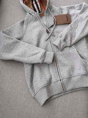 Grå zip hoodie från Burberry - Snygg gråmelerad hoodie från Burberry med dragkedja och klassiskt rutigt foder i huvan. Tröjan har fickor framtill, ribbade muddar och justerbar dragsko i huvan. Perfekt för en chill och stilren look.
