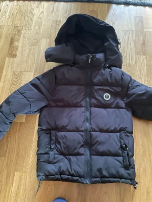 Svart dunjacka med huva från Trapstar - Säljer en svart dunjacka från Trapstar med stor huva och dragkedja framtill. Jackan har två fickor med knappar, Trapstar-logga på bröstet och broderad text på ryggen. Puffig modell som håller dig varm under kalla dagar.