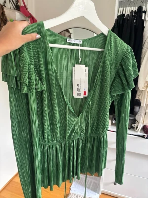 Grön plisserad blus från Zara - Trendig grön plisserad blus från Zara med djup V-ringning och volangdetaljer vid axlarna. Blusen har långa ärmar och ett band i midjan för en snygg siluett. Perfekt för dig som vill sticka ut med färg och struktur.