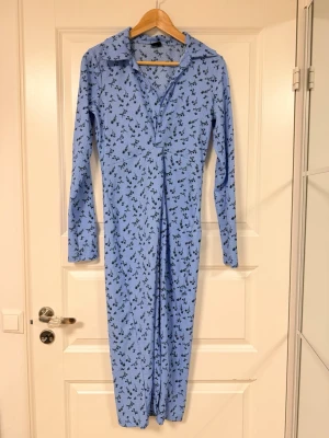 Blå långklänning med blommigt mönster - Långärmad blå långklänning med svart blommigt mönster och krage. Klänningen har v-ringning och är tillverkad i ett mjukt, stretchigt material som ger en slim passform. Perfekt för dig som gillar en feminin och trendig stil.