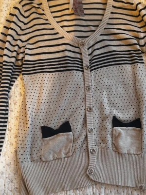 Beige kofta med svarta ränder & rosetter - Beige kofta med svarta horisontella ränder upptill och små svarta prickar nedtill. Två fickor framtill med svarta rosetter som detalj. Djup v-ringning, knäppning med silvriga knappar och ribbade muddar. Långärmad och i mjukt, tunt stickat material. Plus frakt 