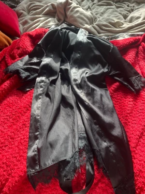 Svart satin morgonrock med spetsdetaljer - Snygg svart morgonrock i glansigt satinmaterial med eleganta spetsdetaljer vid ärmslut och nederkant. Lång modell med tillhörande knytskärp i midjan. Perfekt för mysiga hemmakvällar och en lyxig känsla.