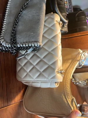 Beige quiltad axelremsväska - Snygg beige axelremsväska i quiltat mönster med silverfärgade detaljer och kedjerem. Väskan har en klassisk rektangulär form och stängs med vridlås. Perfekt för dig som gillar stilrena accessoarer med en lyxig känsla.