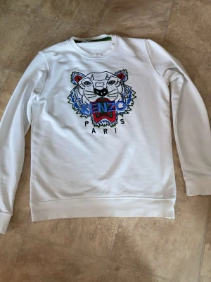 Vit sweatshirt med tigertryck Kenzo - Vit sweatshirt från Kenzo med det ikoniska broderade tigerhuvudet i flera färger på bröstet och texten 'KENZO PARIS' under. Tröjan har rund halsringning, långa ärmar och ribbade muddar. Perfekt för dig som gillar streetwear och vill sticka ut.