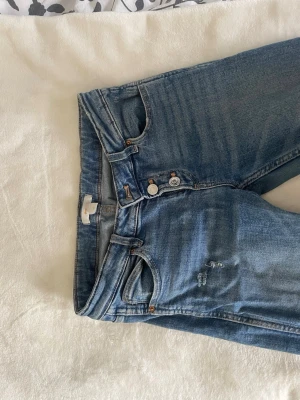 Bootcut jeans gina tricot  - Säljer ett par ljusblå jeans med bootcut och lågmidja. Jeansen har klassisk femficksmodell, slitningar på ena fickan och ett hål på benet samt knappgylf. Perfekta för en avslappnad och trendig look. Säljer då dom är för korta 