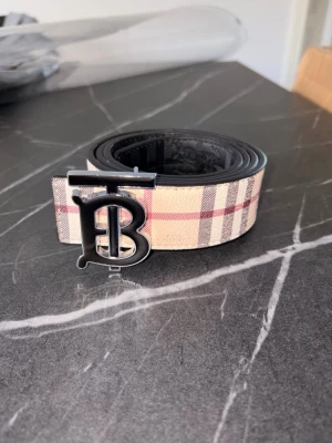 Beige och svart bälte med rutor - Snyggt bälte i beige och svart med rutigt mönster och en stor metallspänne med bokstäver. Perfekt accessoar för att lyfta din outfit och ge en trendig touch. Bältet har klassiska färger och känns riktigt stilrent.